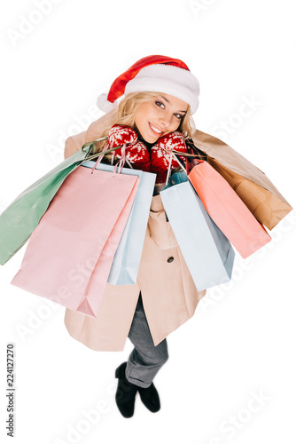high angle view of happy young woman in santa hat and mittens holding shopping bags and smiling at camera isolated on white