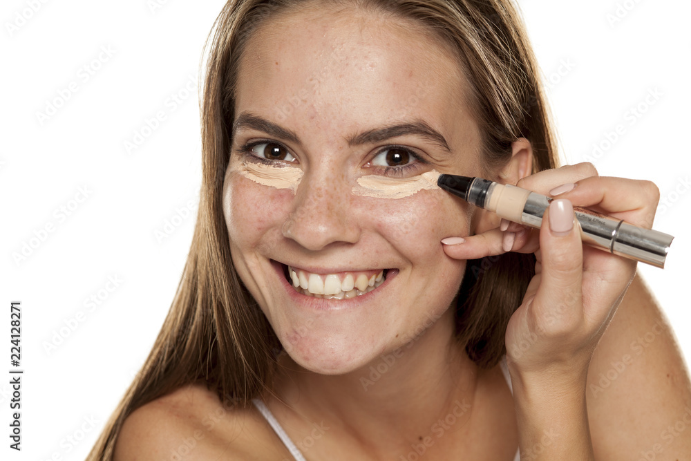 young beautiful girl applying concealer under her eyes on white backgeound