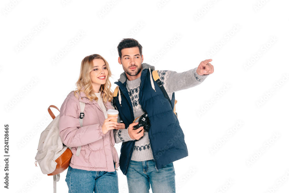 young couple of tourists with backpacks holding coffee to go and camera while pointing and looking away isolated on white young couple of tourists with backpacks holding coffee to go and camera while pointing and looking away isolated on white