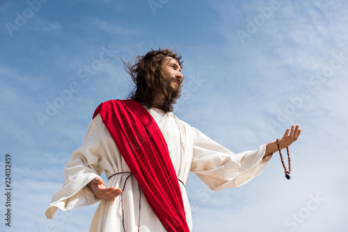 Принти на полотні low angle view of Jesus in robe holding rosary and reaching hand against blue sk