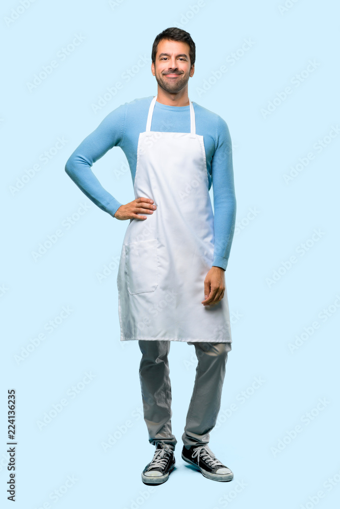 Full body of Man wearing an apron posing with arms at hip and smiling on blue background Full body of Man wearing an apron posing with arms at hip and smiling on blue background