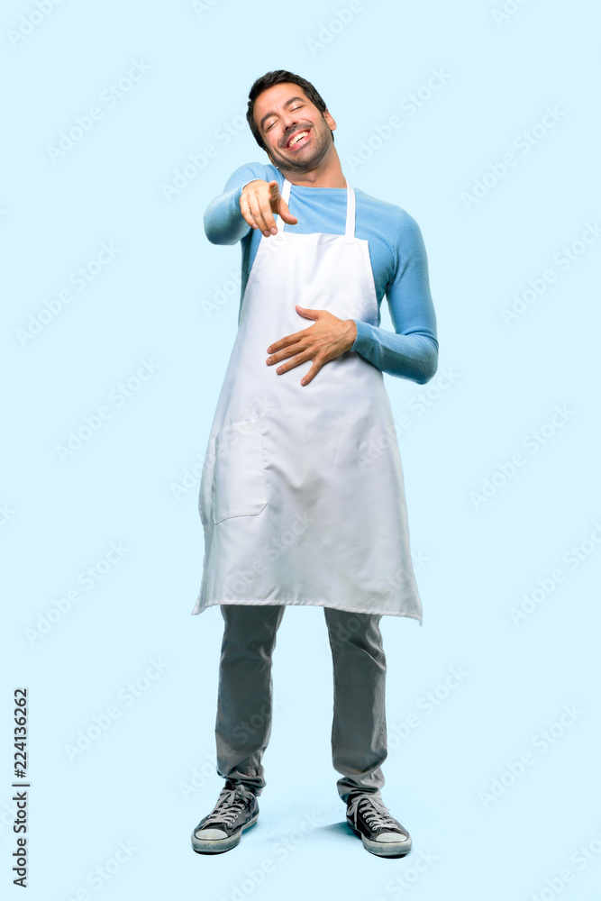 Full body of Man wearing an apron pointing with finger at someone and laughing a lot on blue background Full body of Man wearing an apron pointing with finger at someone and laughing a lot on blue background