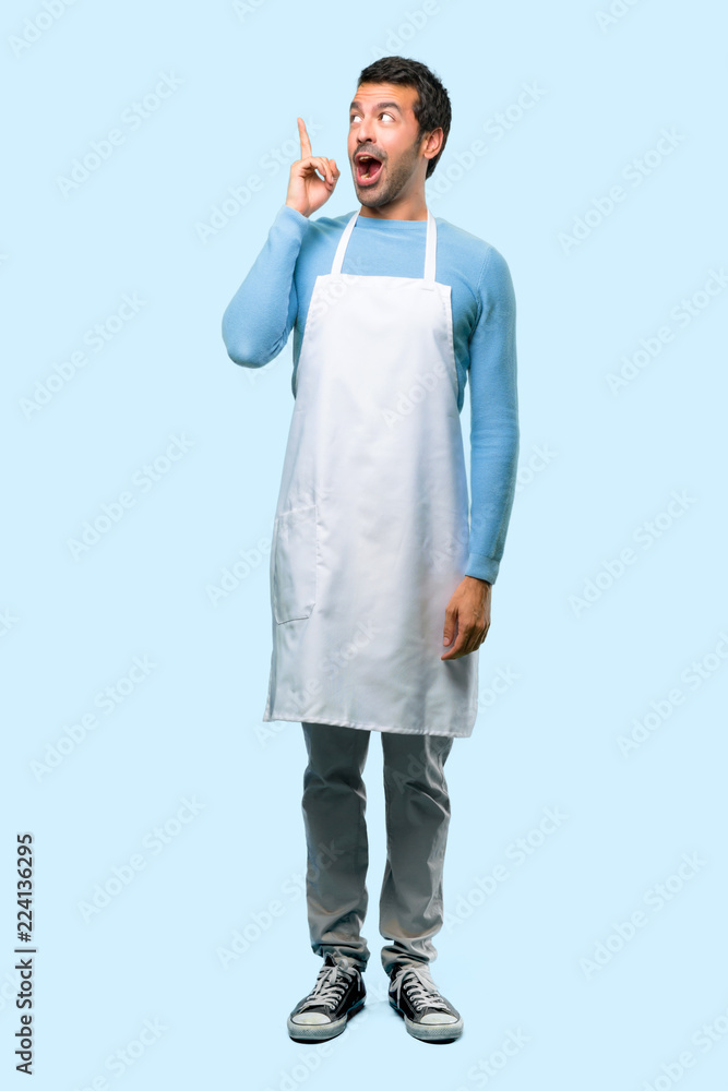 Full body of Man wearing an apron intending to realizes the solution while lifting a finger up on blue background Full body of Man wearing an apron intending to realizes the solution while lifting a finger up on blue background