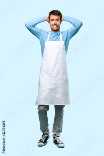 Full body of Man wearing an apron takes hands on head because has migraine on blue background