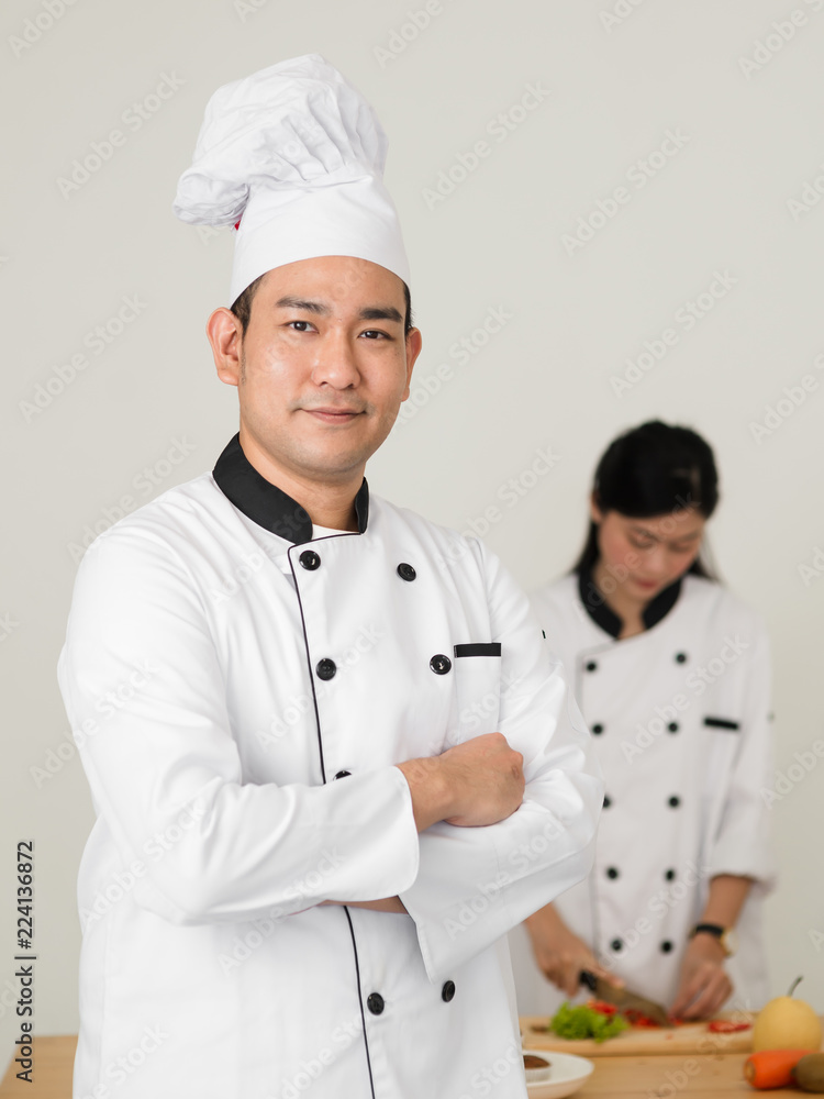 Confident Asian chef looking at camera with his helper background
