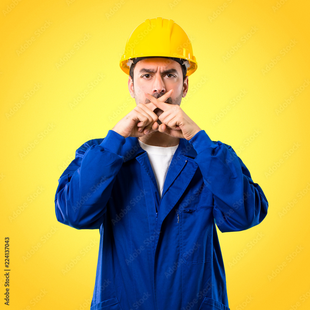 Young workman with helmet showing a sign of closing mouth and silence gesture on yellow background Young workman with helmet showing a sign of closing mouth and silence gesture on yellow background