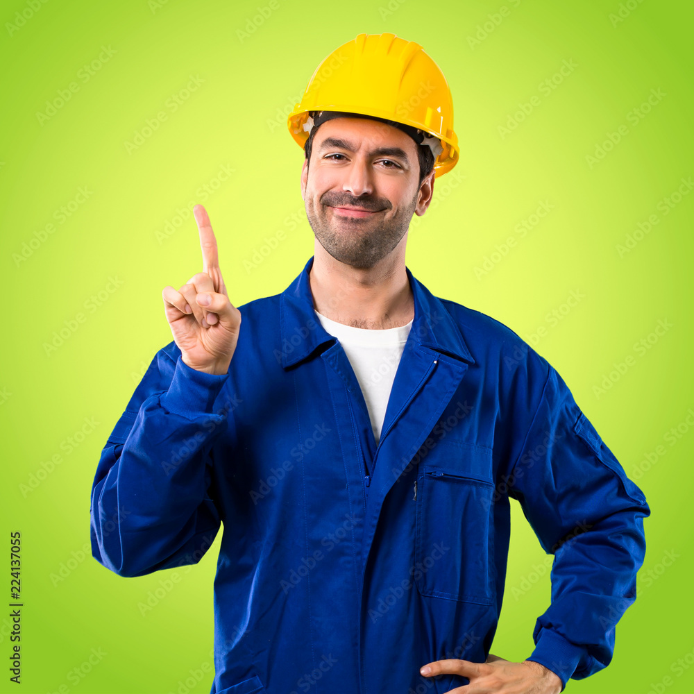 Young workman with helmet showing and lifting a finger in sign of the best on green background Young workman with helmet showing and lifting a finger in sign of the best on green background