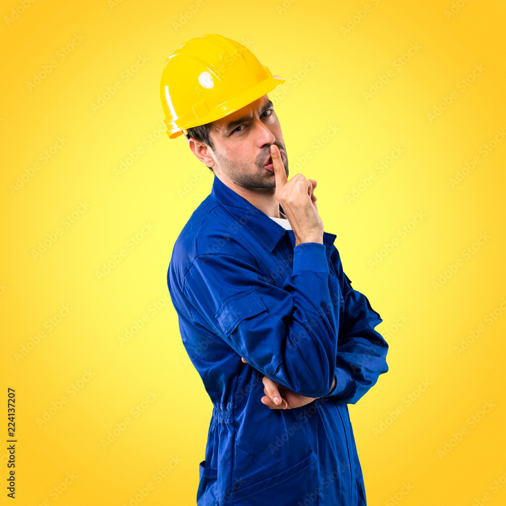 Young workman with helmet showing a sign of closing mouth and silence gesture putting finger in mouth on yellow background