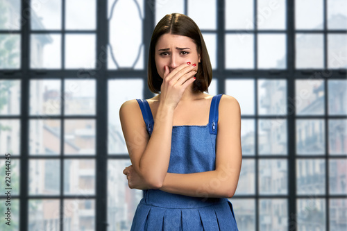 Girl is crying on blurred background. Portrait of young upset woman covering mouth with hand and crying on abstact window background. Human negative expressions.