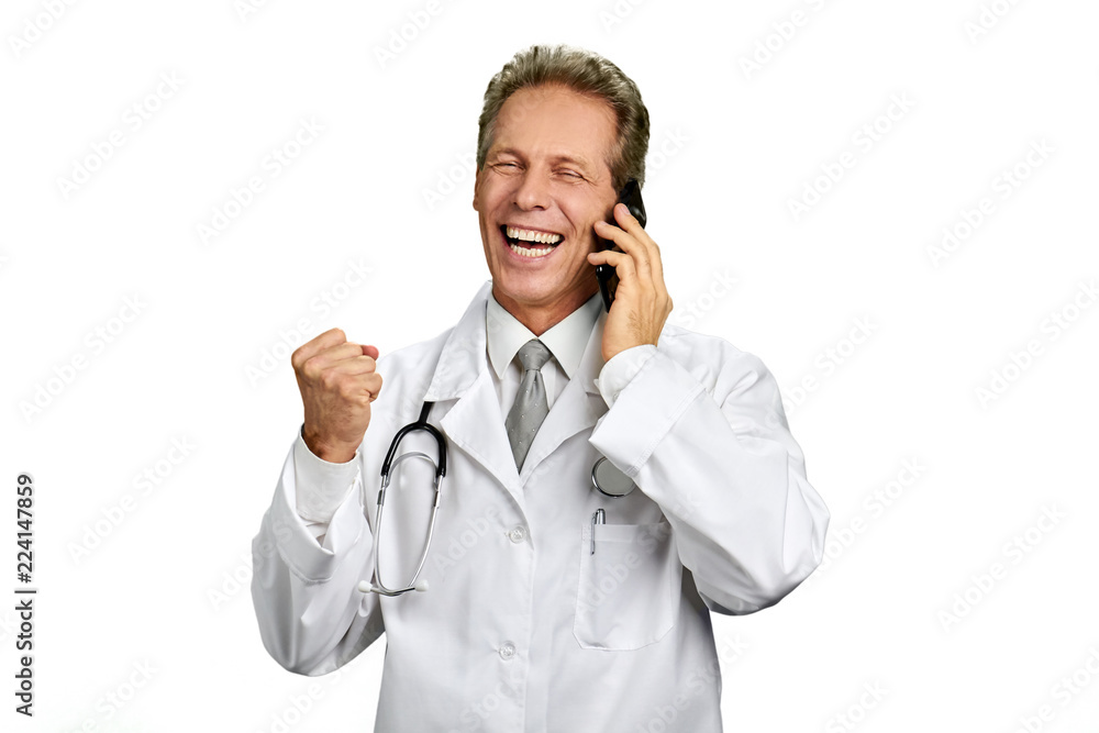 Joyful male doctor talking on phone. Happy male practitioner taking on cell phone and clenched his fist in excitement over white background. Gesture of victory. Joyful male doctor talking on phone. Happy male practitioner taking on cell phone and clenched his fist in excitement over white background. Gesture of victory.