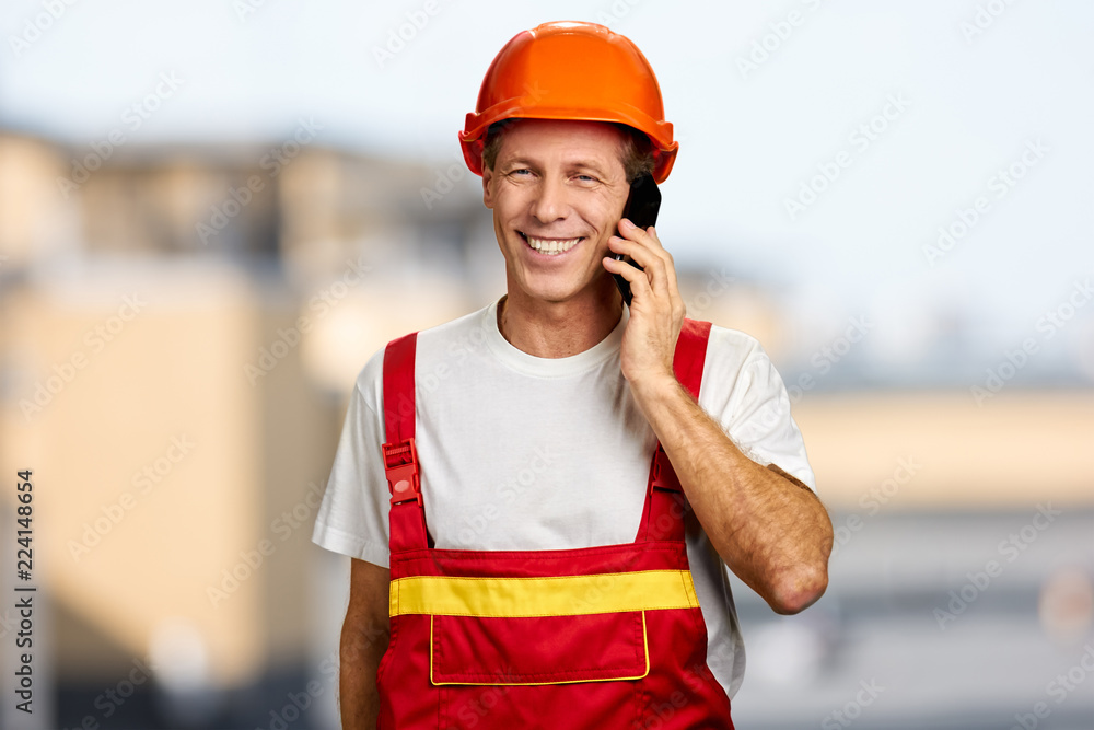 Happy worker in hard hat talking on phone. Cheerful foreman in uniform talking on telephone on blurred background. Happy worker in hard hat talking on phone. Cheerful foreman in uniform talking on telephone on blurred background.