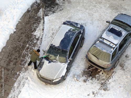 The man is cleaning the windshield from the snow in order to get out onto the streets of the city. A view from above of a man who cleans his car from an icy captivity. Parking on the street.