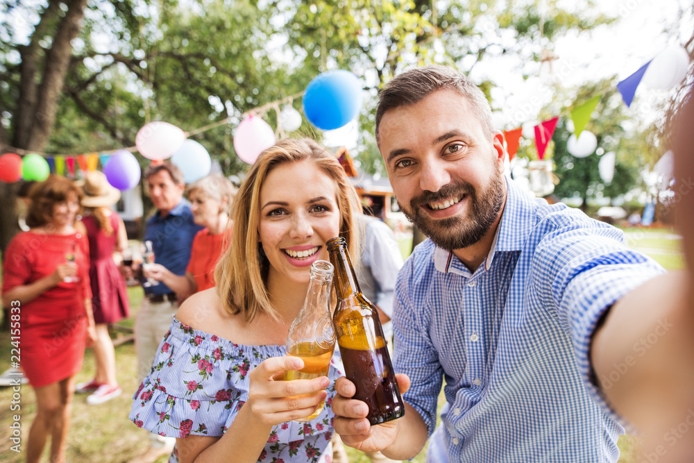 Obraz premium Young couple taking selfie at a party outside in the backyard, clinking bottles.