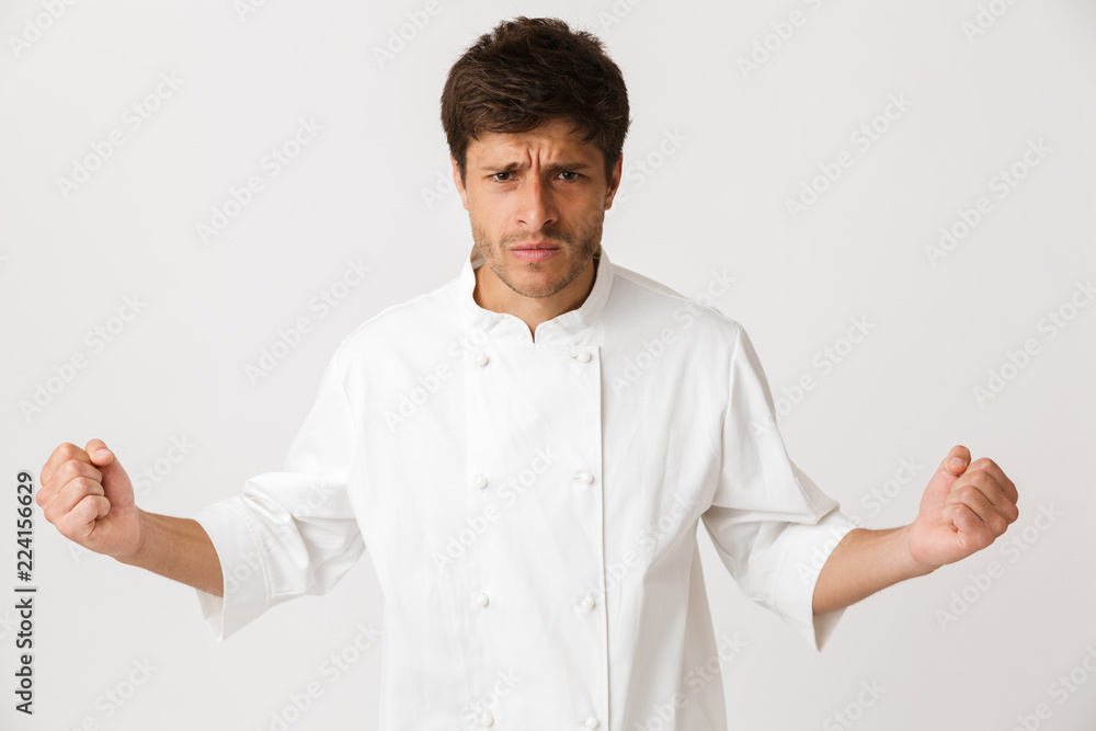 Angry young chef man standing isolated over white wall background. Angry young chef man standing isolated over white wall background.