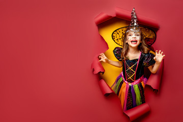 Happy Halloween. Laughing funny child girl in a witch costume of halloween looking, smiling and scares through a hole of red, yellow paper background. Copyspace
