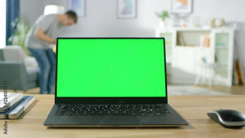 Modern Laptop with Green Mock-up Screen Display Standing on the Desk in the Cozy Living Room. Man with Mobile Phone Sits on a Chair to Rest. Modern Laptop with Green Mock-up Screen Display Standing on the Desk in the Cozy Living Room. Man with Mobile Phone Sits on a Chair to Rest.