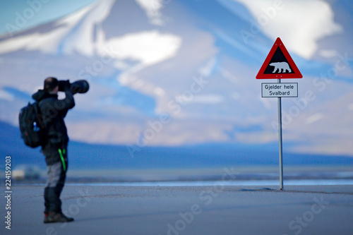 Photographer with big lens and road traffic sign with Polar bear. “Gjelder Hele Svalbard” means “Over All of Svalbard (watch out for polar bears)”. Man on the road with snowy mountain, Svalbard. photo