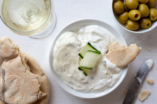Tzatziki in white ceramic bowl with a cucumber slice and a piece of pita bread next to pita bread, olives and a glass of white wine from above.