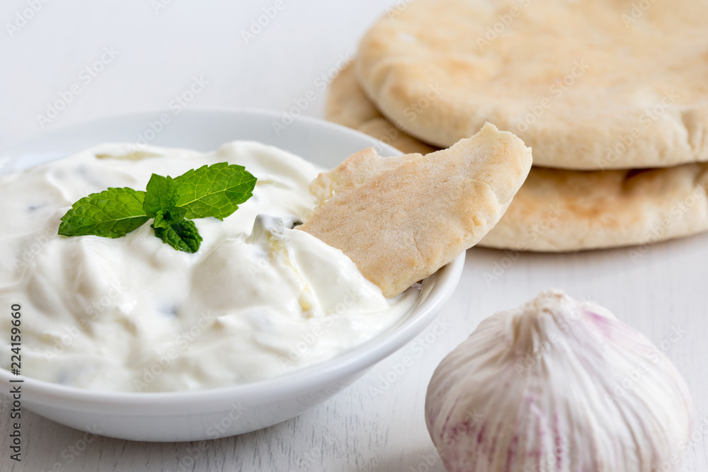 Tzatziki in white ceramic bowl with mint leaf garnish and a piece of pita bread next to pita bread and garlic bulb. Tzatziki in white ceramic bowl with mint leaf garnish and a piece of pita bread next to pita bread and garlic bulb.