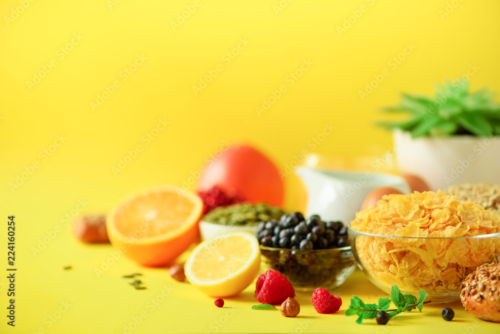 Corn cereal, muesli, milk, berries, orange juice, yogurt, boiled egg, nuts, fruits, banana, peach for breakfast on yellow background. Copy space. Vegan and vegetarian concept. Corn cereal, muesli, milk, berries, orange juice, yogurt, boiled egg, nuts, fruits, banana, peach for breakfast on yellow background. Copy space. Vegan and vegetarian concept.