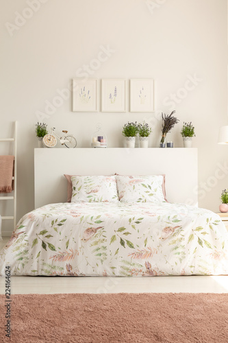 Pink carpet and posters in bright bedroom interior with plants above bed with cushions. Real photo