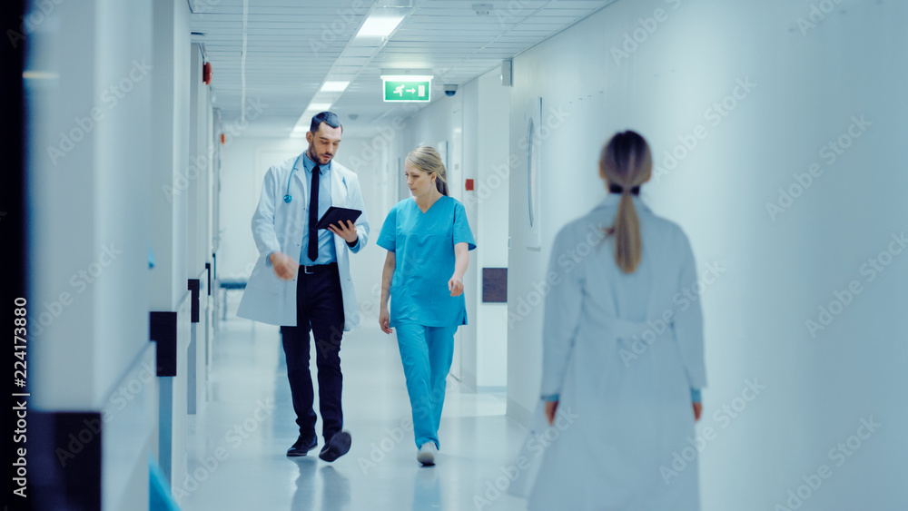 Female Surgeon and Doctor Walk Through Hospital Hallway, They Consult Digital Tablet Computer while Talking about Patient's Health. Modern Bright Hospital with Professional Staff. Female Surgeon and Doctor Walk Through Hospital Hallway, They Consult Digital Tablet Computer while Talking about Patient's Health. Modern Bright Hospital with Professional Staff.