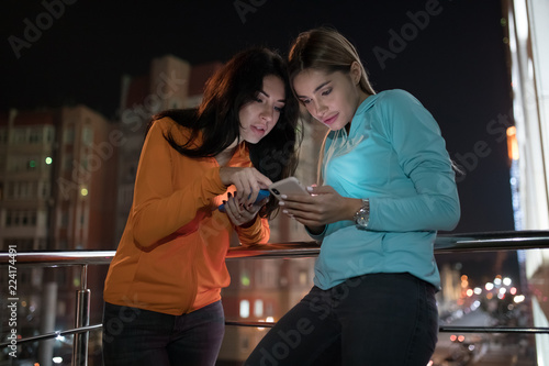 Young women friends looking amazedly at smartphone in the balcony terrace, night city background photo