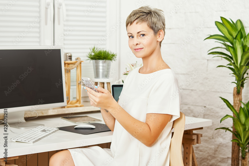 beautiful business woman in a white dress sitting at a desk with a phone in hand and works on the computer in a home office beautiful business woman in a white dress sitting at a desk with a phone in hand and works on the computer in a home office