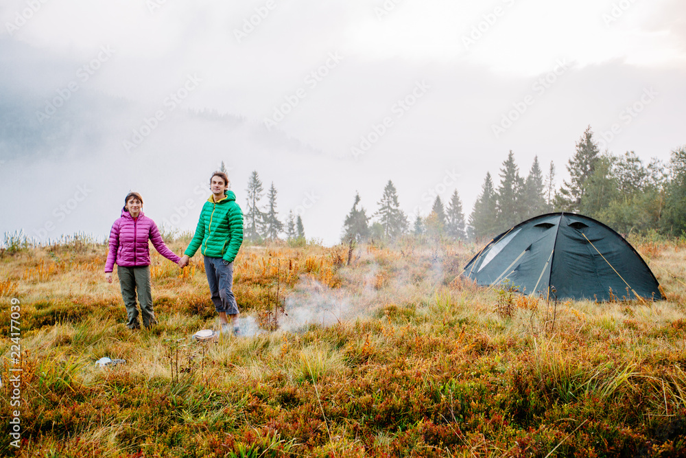 Autumn nature lifestyle happy man and woman relaxing outdoors on camping travel park with frog on background. Hiking couple wearing green and purple down jacket enjoying fall season scenic landscape.