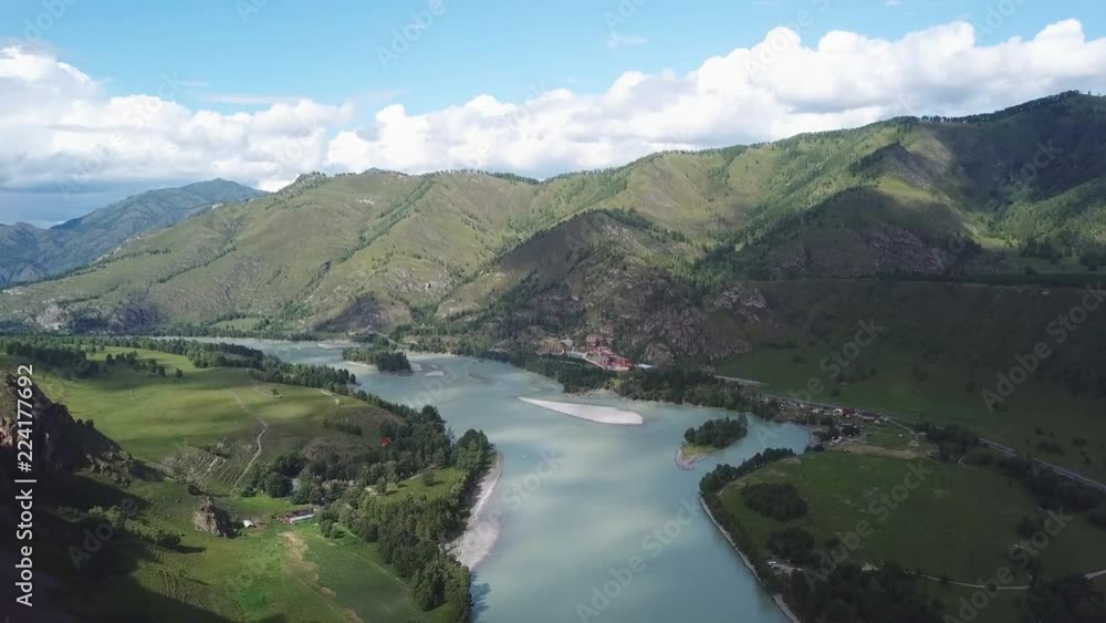Aerial view above the turquoise river Katun among the trees and meadows in the mountains of the Altai under white clouds and a blue sunny sky flying towards the tourist base with houses and buildings Aerial view above the turquoise river Katun among the trees and meadows in the mountains of the Altai under white clouds and a blue sunny sky flying towards the tourist base with houses and buildings