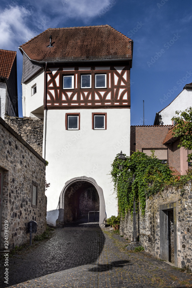 Germany, Rhine-Main area, Hanau, Steinheim: Ancient white half-timbered main gate in the city center of the German town with cobblestone road, old city wall and blue sky - concept travel fortification