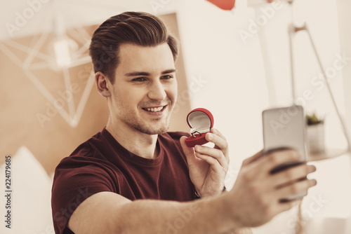 Selfie time. Smiling excited man feeling nervous while making selfie with expensive wedding ring for his sweetheart