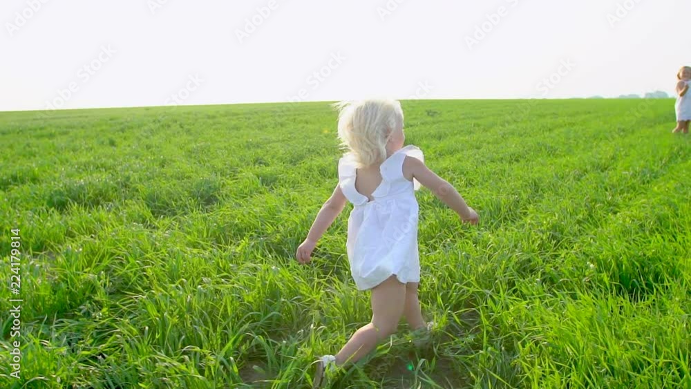 Blond little cute girl in white dress running on the green field to sister and smiling. Sisters, childs, kids running in garden and laughing. Concept of beauty, nature, love, health, childhood, happy Blond little cute girl in white dress running on the green field to sister and smiling. Sisters, childs, kids running in garden and laughing. Concept of beauty, nature, love, health, childhood, happy