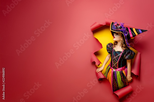 Happy Halloween. Laughing funny child girl in a witch costume of halloween looking to the side through a hole of red and yellow paper background. Copyspace