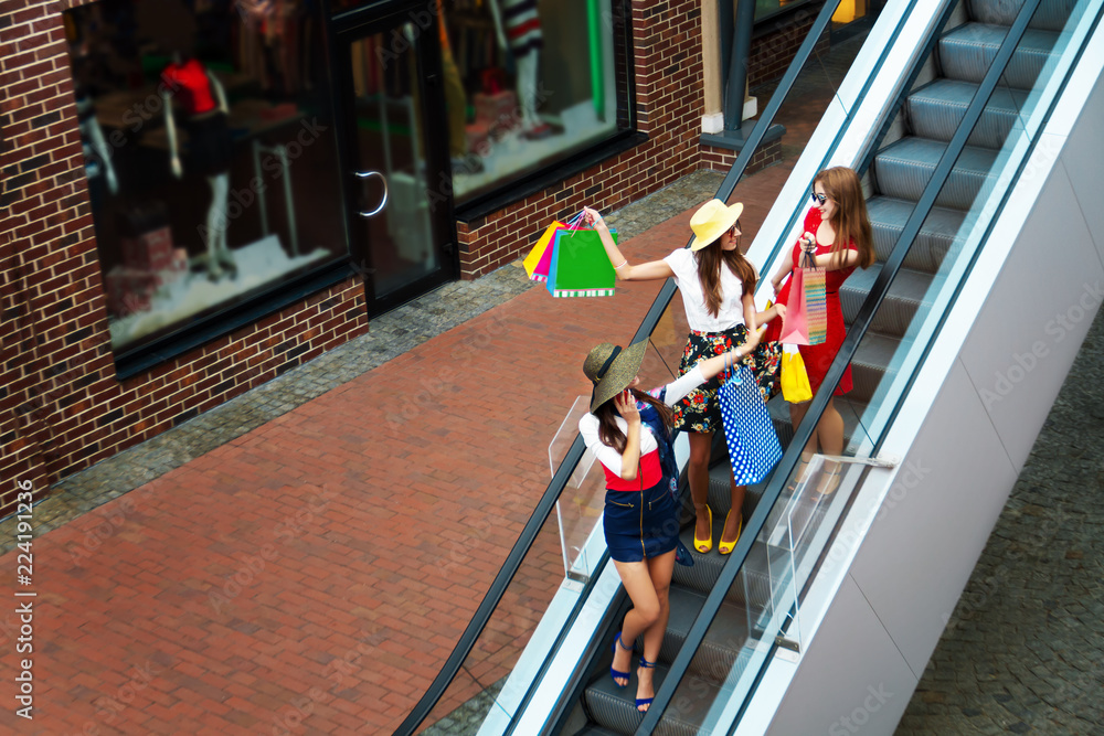 Obraz premium Pretty happy bright women female girls friends in colorful dresses, hats and high heels with shopping bags walking at street after shopping in shopping mall in summer. Shop sales, black Friday.