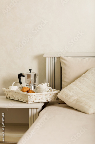Breakfast in bed. On a white wicker tray there is a coffee maker, coffee white cups and croissants.