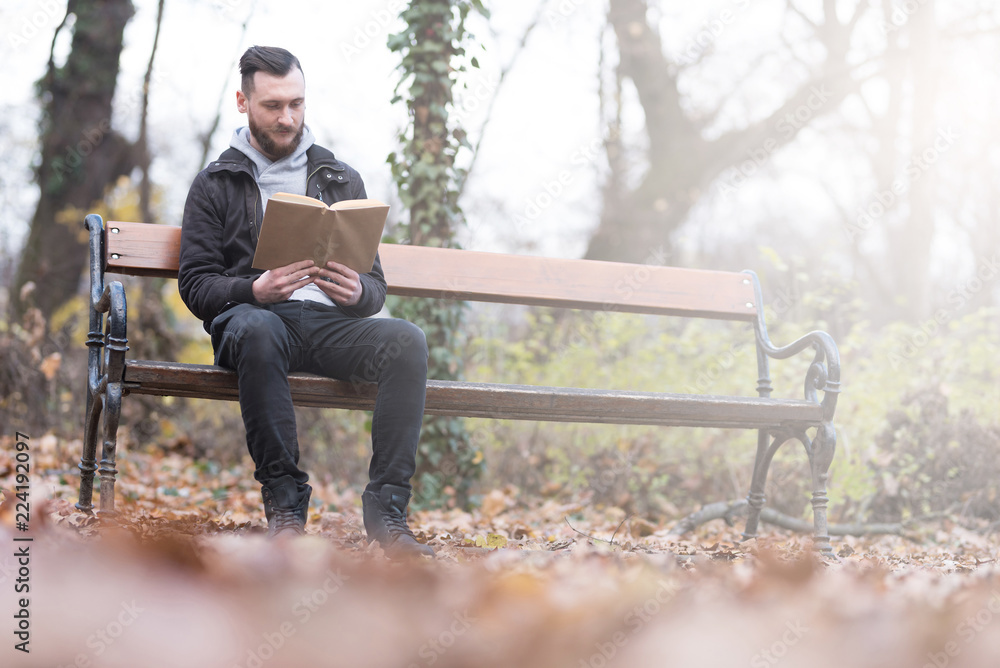 A handsome young hipster man sitting on a bench and reading a book in the park. A handsome young hipster man sitting on a bench and reading a book in the park.