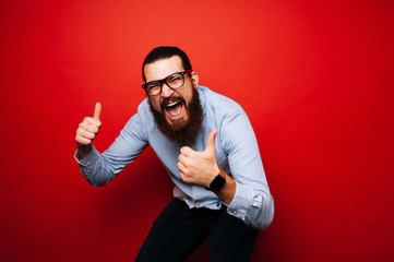 Cheerful bearded hipster man in casual with eyeglasses jump over red background and showing thumbs up and wearing smartwatch