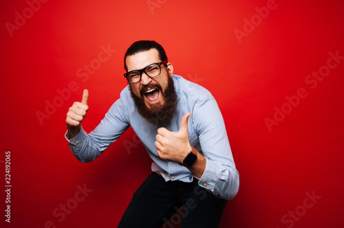 Cheerful bearded hipster man in casual with eyeglasses jump over red background and showing thumbs up and wearing smartwatch