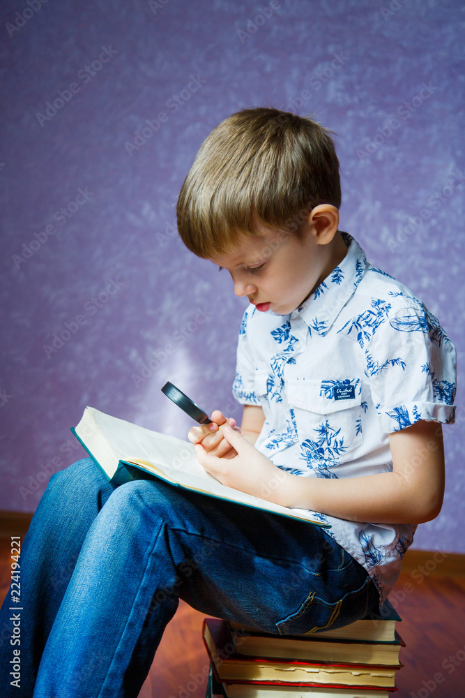 Fototapeta premium Little boy in a light shirt sits on a pile of books with a green book in his hands and looks through a magnifying glass