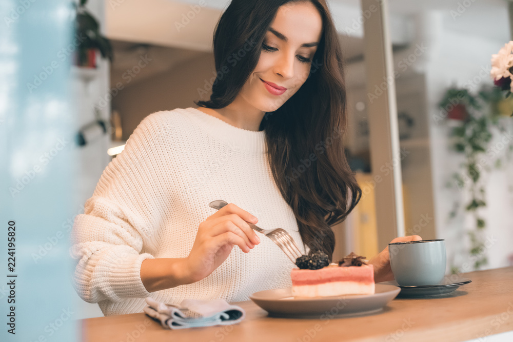 selective focus of young woman with coffee cup eating cheesecake at table in cafe selective focus of young woman with coffee cup eating cheesecake at table in cafe