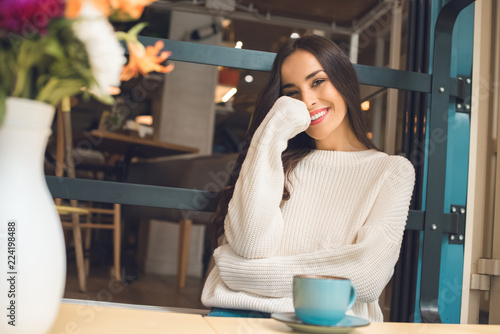 happy attractive young woman looking at camera at table with coffee cup in cafe