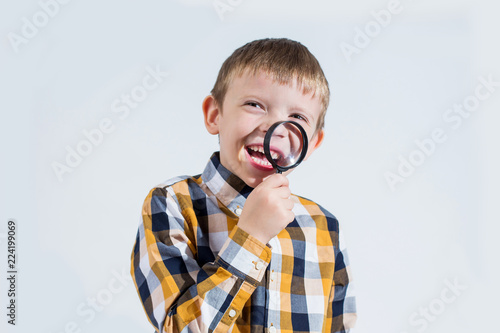 Little boy in a plaid shirt on a white background laughs gaily and looks with one eye through a magnifier