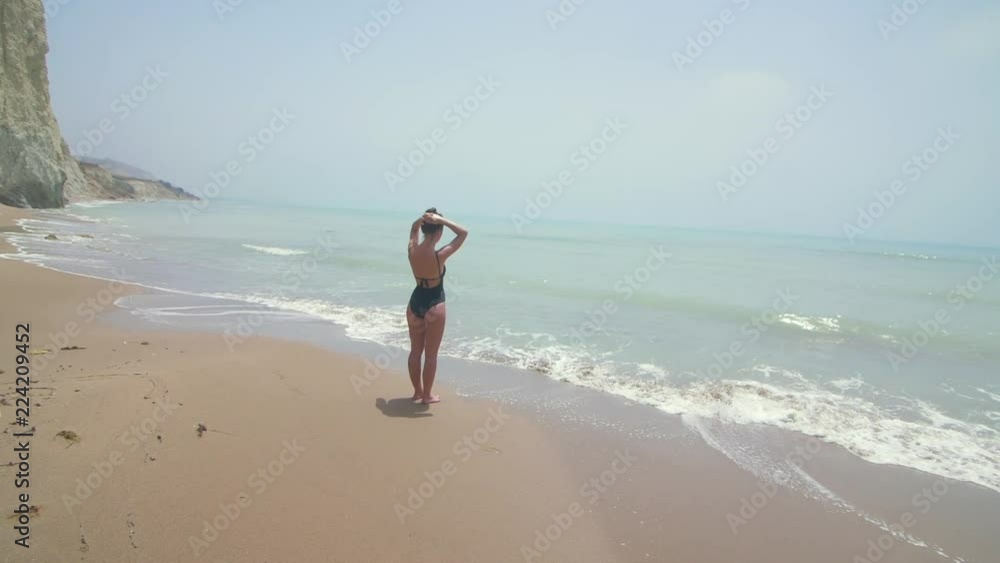Happy young girl, sea and summer holidays. Beautiful brunette model standing on tropical beach. Woman dressed in black swimsuit and hat. Enjoys her vacation. Slow motion Happy young girl, sea and summer holidays. Beautiful brunette model standing on tropical beach. Woman dressed in black swimsuit and hat. Enjoys her vacation. Slow motion