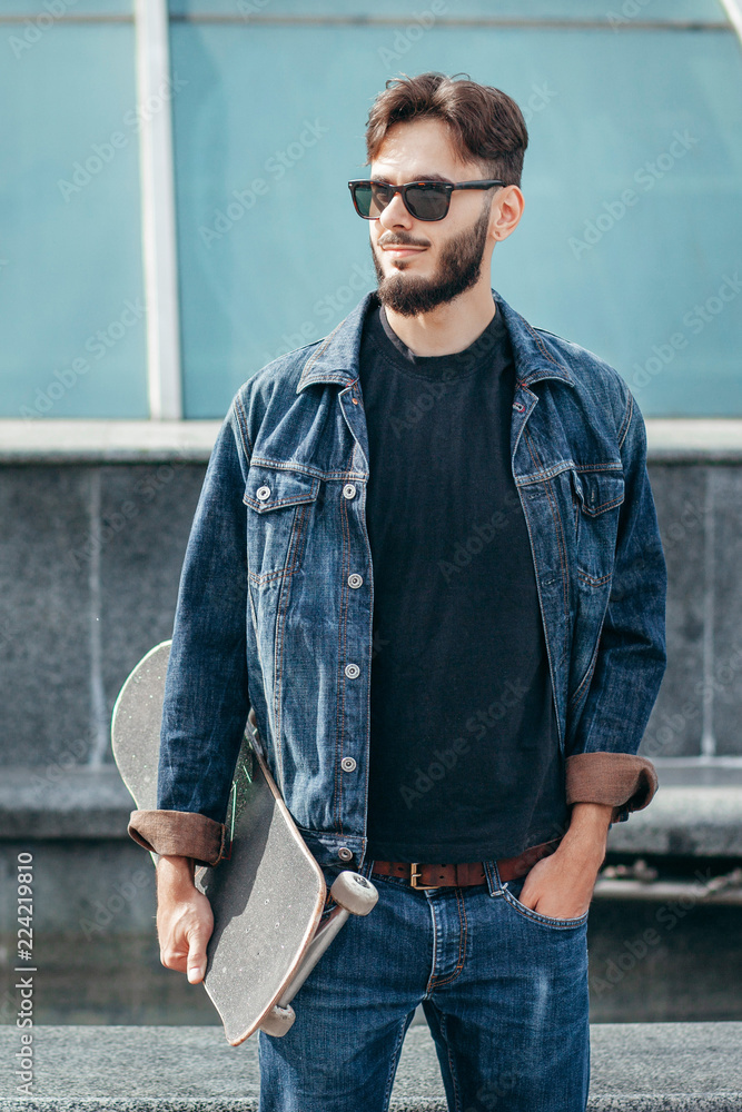 oung serious guy in black T-shirt looking in camera holding skateboard in hand with modern background oung serious guy in black T-shirt looking in camera holding skateboard in hand with modern background