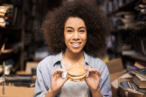 Young woman holding in hands fast food hamburger, american unhealthy calories meal, books on background, fast food break of hungry student preparing exam smiling with grilled cheesburger front view
