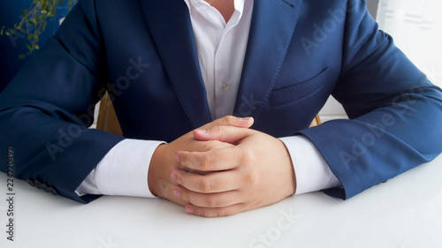 Closeup image of businessman in blue suit with folded hands sitting behind white wooden office desk