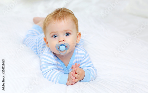 Charming blue-eyed baby 7 month old lies in bed in a striped bodysuit and sucks a pacifier