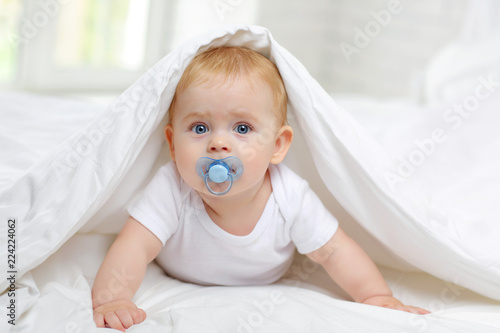 Charming blue-eyed baby 7 month old lies in bed in a white bodysuit, covered with a blanket and sucks a pacifier