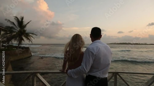 Back view of bride and groom enjoying sunset on tropical beach near balustrade on vacation. Newlyweds hugging and watching ocean after ceremony on honeymoon. Tropical wedding at Maldives luxury spa